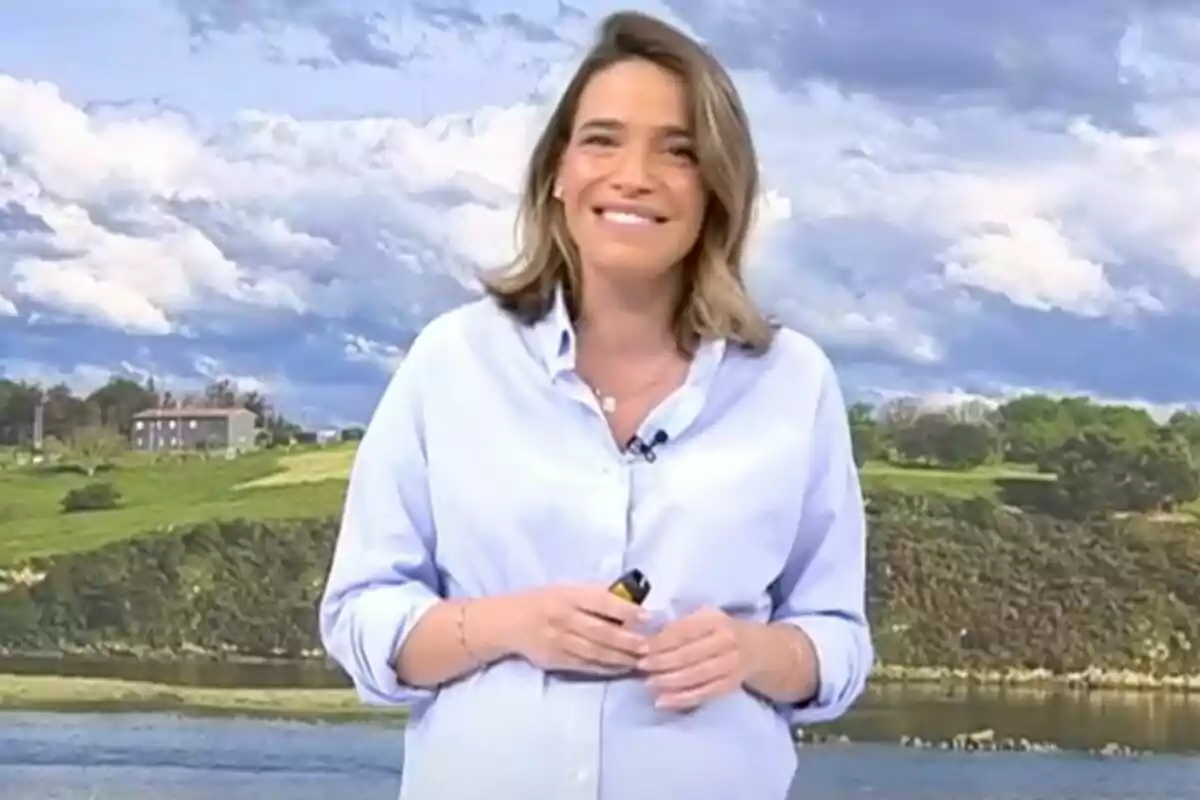 Flora González somrient amb camisa blau clar està dreta davant d'un paisatge amb cel ennuvolat i turons verds a 'Informatius Telecinco'.