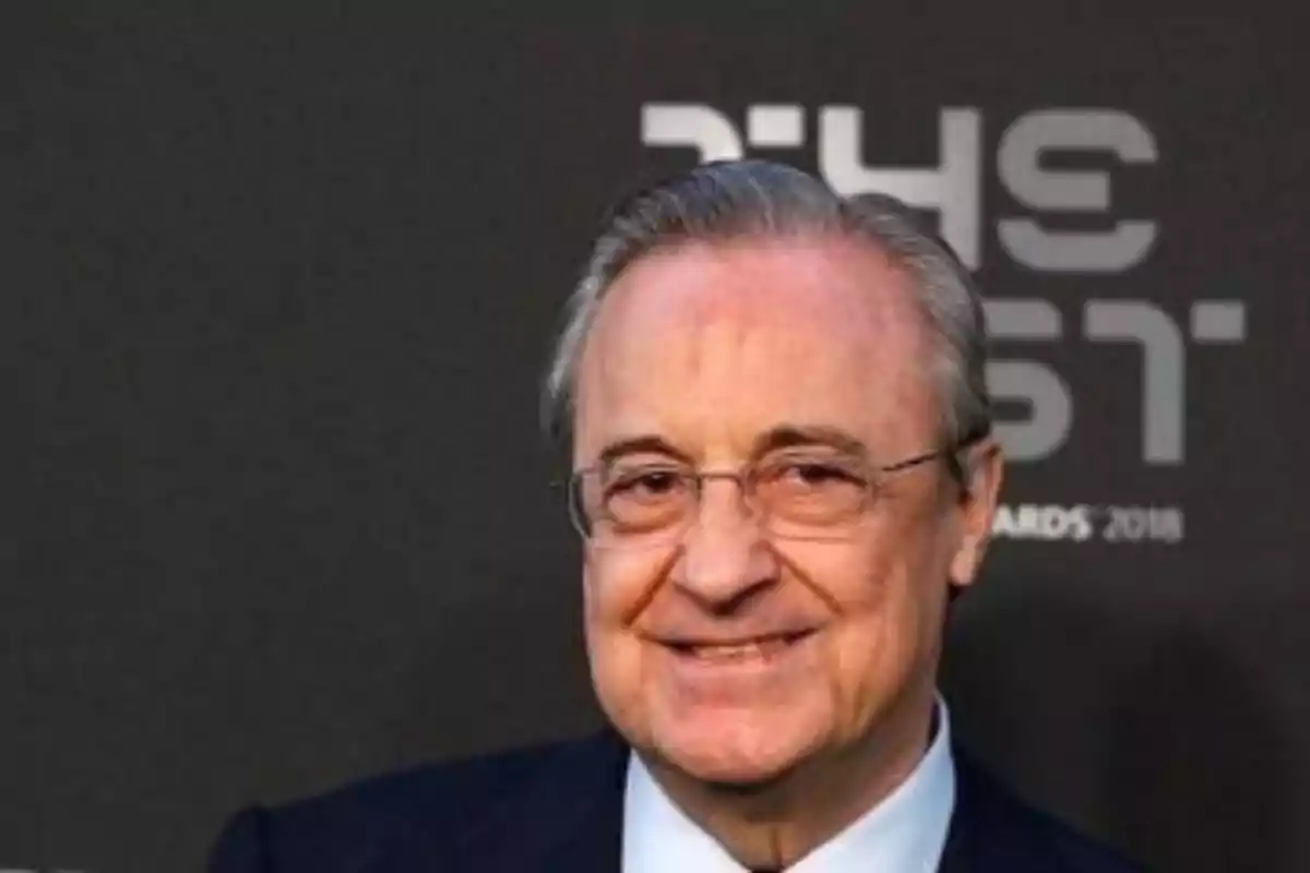 A man with glasses and a suit smiles in front of a dark background with text. A man with glasses and a suit smiles in front of a dark background with text.