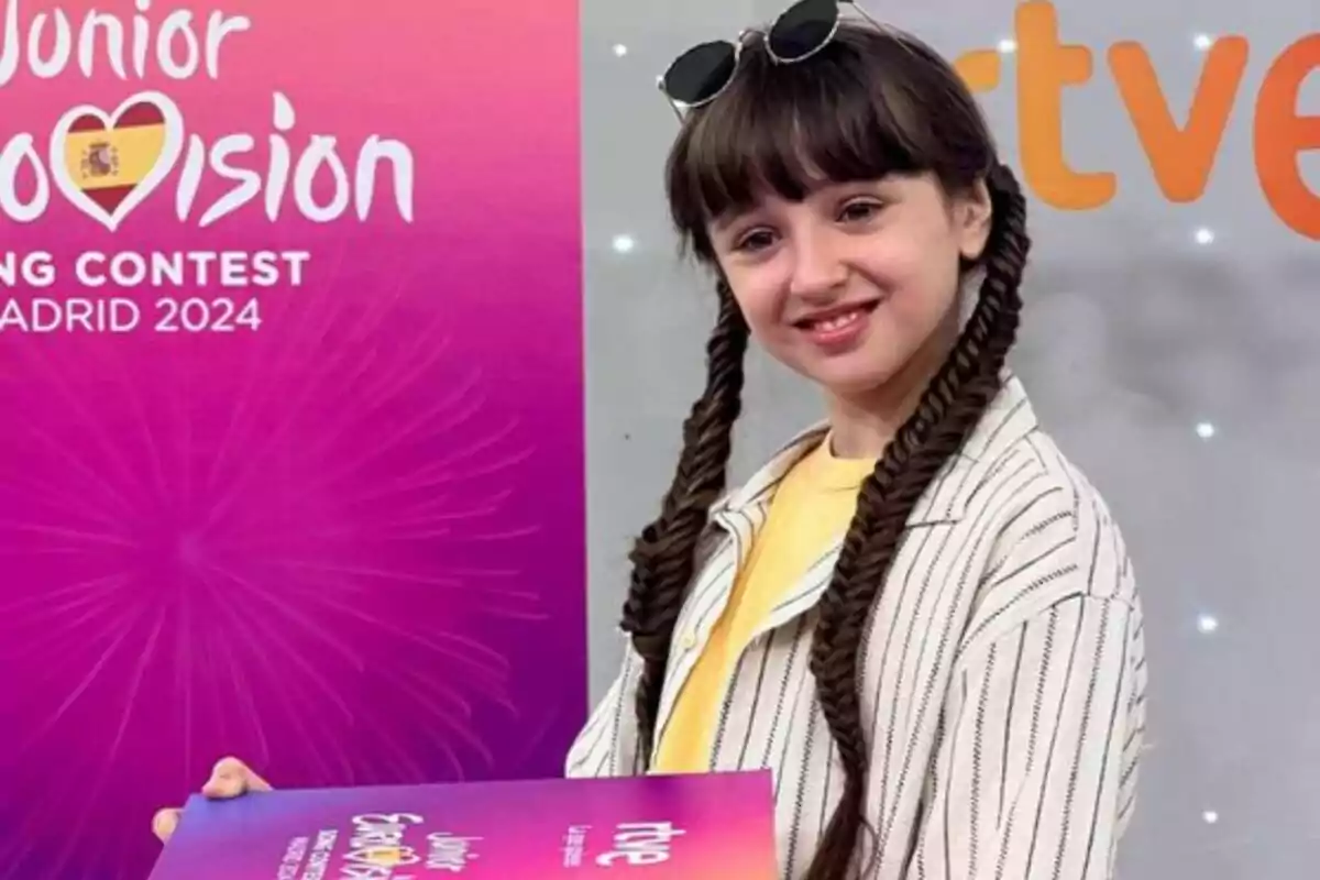 Una nena amb trenes i ulleres de sol al cap sosté un cartell del Junior Eurovision Song Contest Madrid 2024 davant d'un fons rosa amb el logo del concurs.