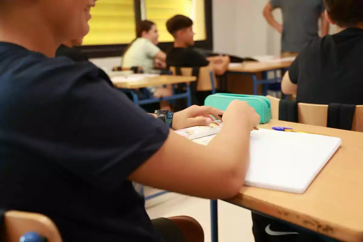 Estudiants asseguts a una aula, alguns prenent notes i altres conversant, amb un estoig de llapis verd sobre l'escriptori.