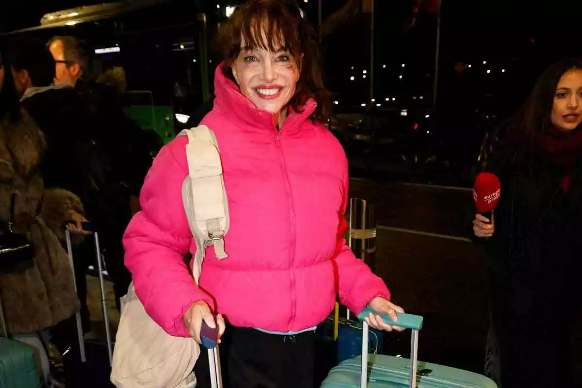 Beatriz Rico in a pink jacket and white backpack smiles while holding a blue suitcase in a crowded place. Beatriz Rico in a pink jacket and white backpack smiles while holding a blue suitcase in a crowded place.