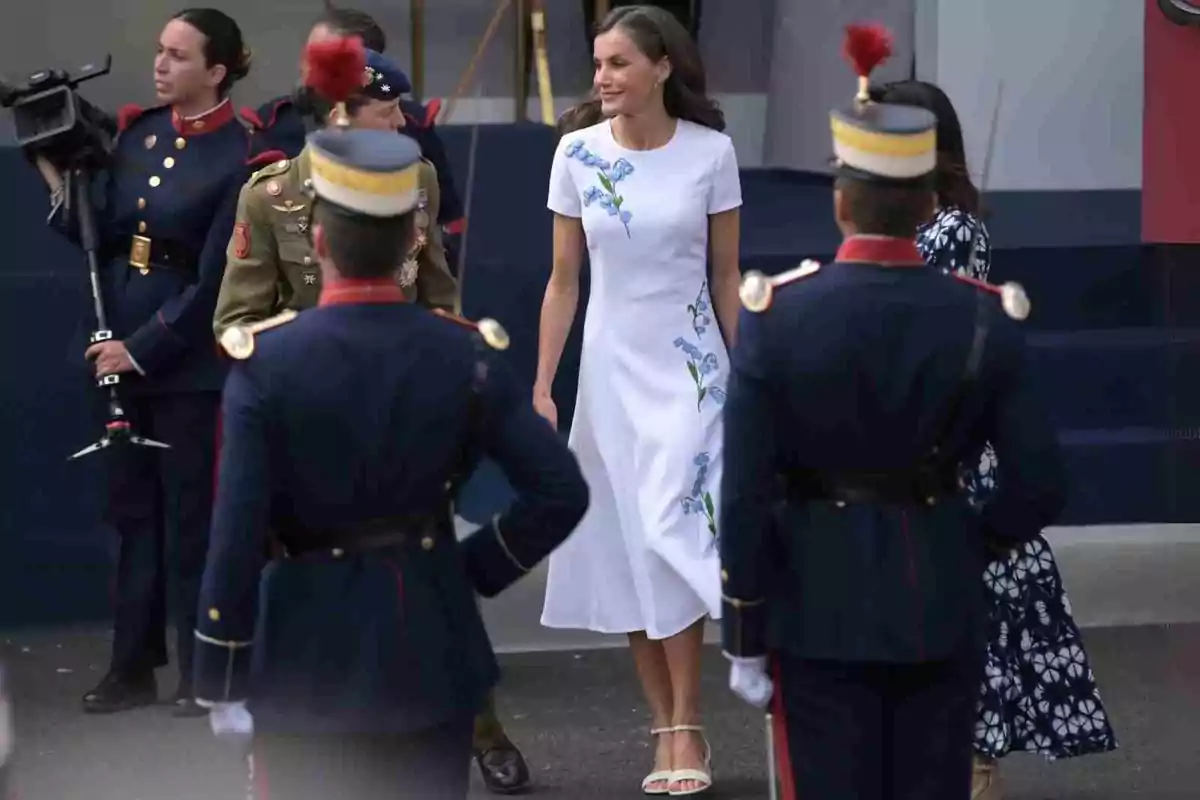 La reina Letícia amb vestit blanc decorat amb flors blaves caminant entre membres de la guàrdia vestits amb uniformes formals La reina Letícia amb vestit blanc decorat amb flors blaves caminant entre membres de la guàrdia vestits amb uniformes formals