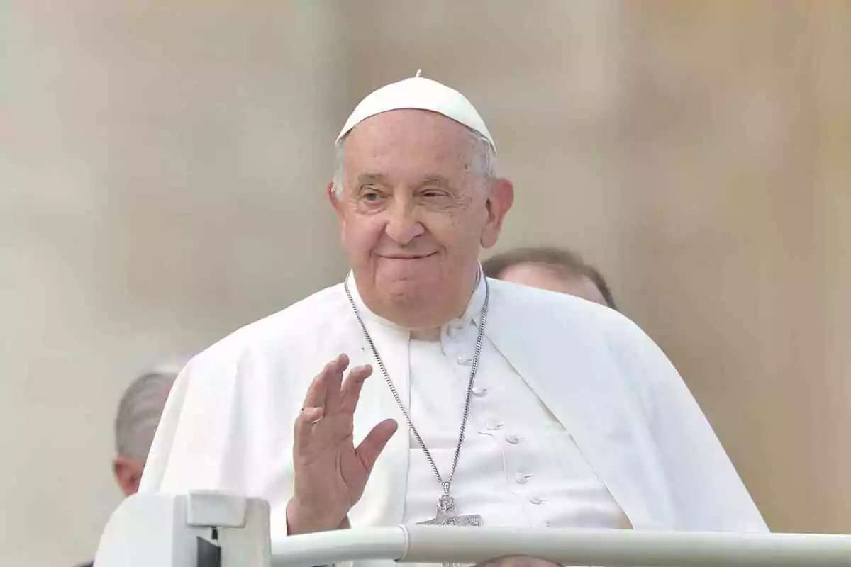 El papa Francisco vestido con vestimenta religiosa blanca, incluyendo un solideo y una cruz colgante, saludando con una mano.