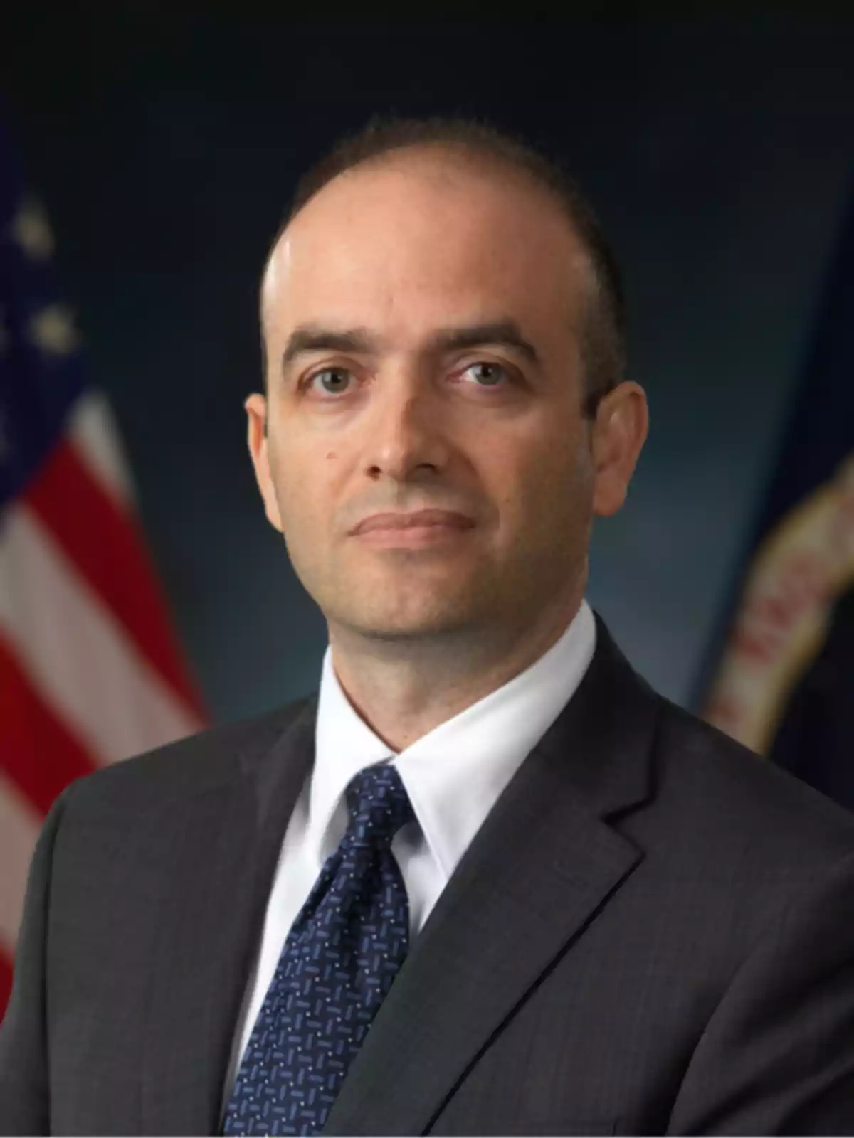 Omar Hatamleh con traje oscuro y corbata azul posa frente a una bandera estadounidense.