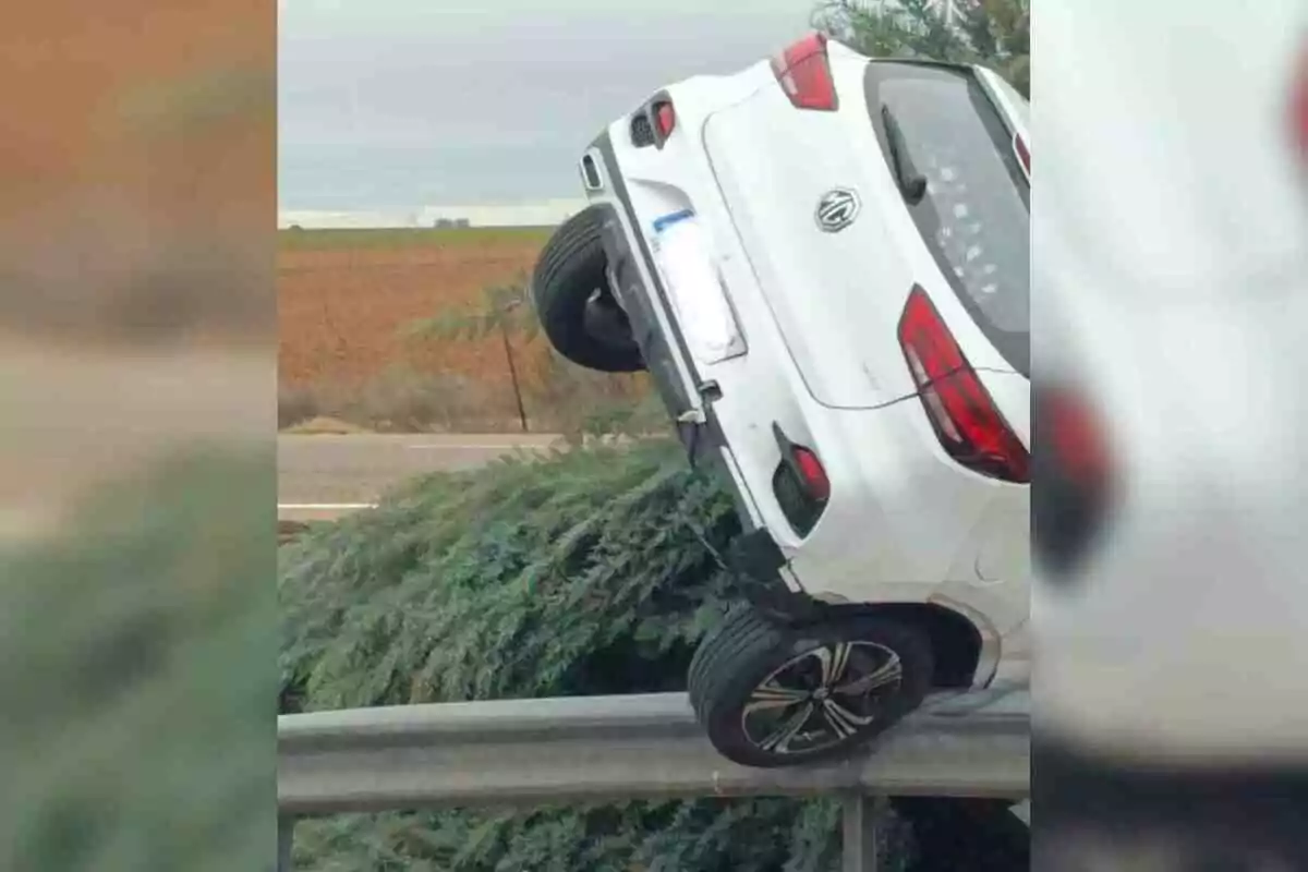 Un coche blanco está inclinado sobre una barrera de seguridad en una carretera rural.