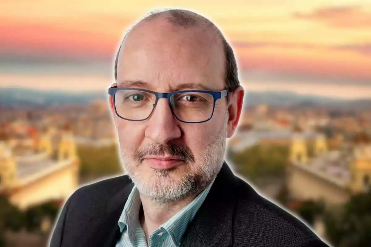 A man with glasses and a beard in front of a blurred urban background at sunset.