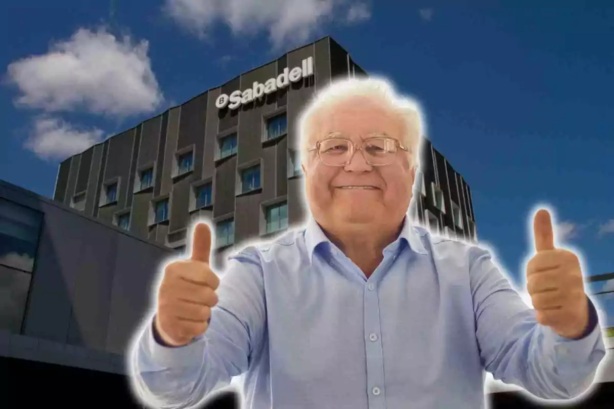 Un home gran somrient amb ulleres i camisa blava aixeca ambdós polzes davant d'un edifici amb el logotip de Sabadell en un dia assolellat.