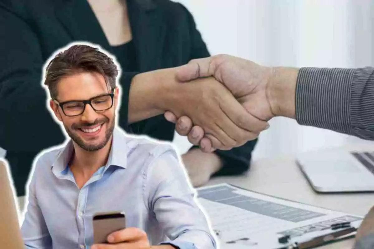 Un home somrient amb ulleres i barba mira el telèfon mòbil mentre dues persones es donen la mà al fons, suggerint un acord o col·laboració.