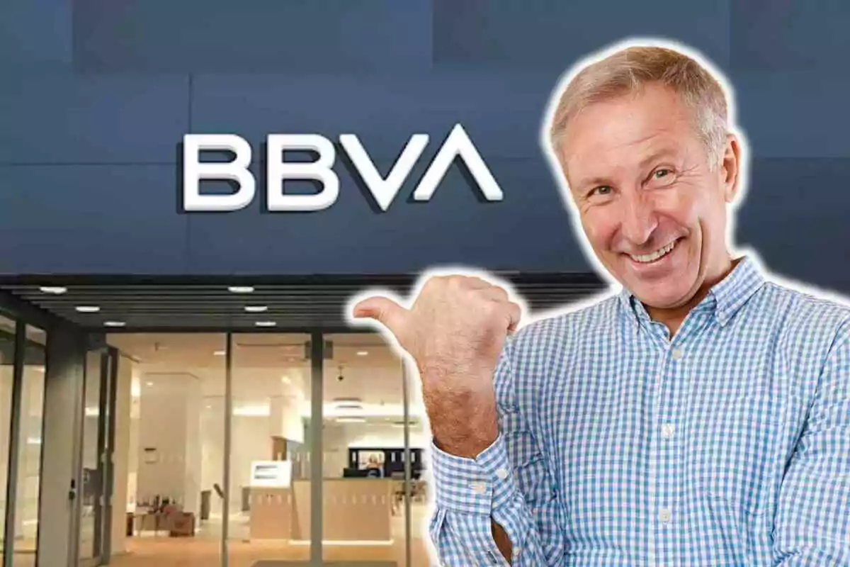 Un home somrient amb camisa a quadres blaus i blancs assenyala amb el polze cap a una sucursal del banc BBVA.