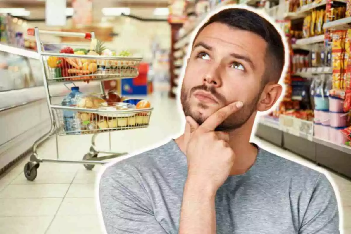 Hombre pensativo en un supermercado con un carrito de compras lleno de alimentos.