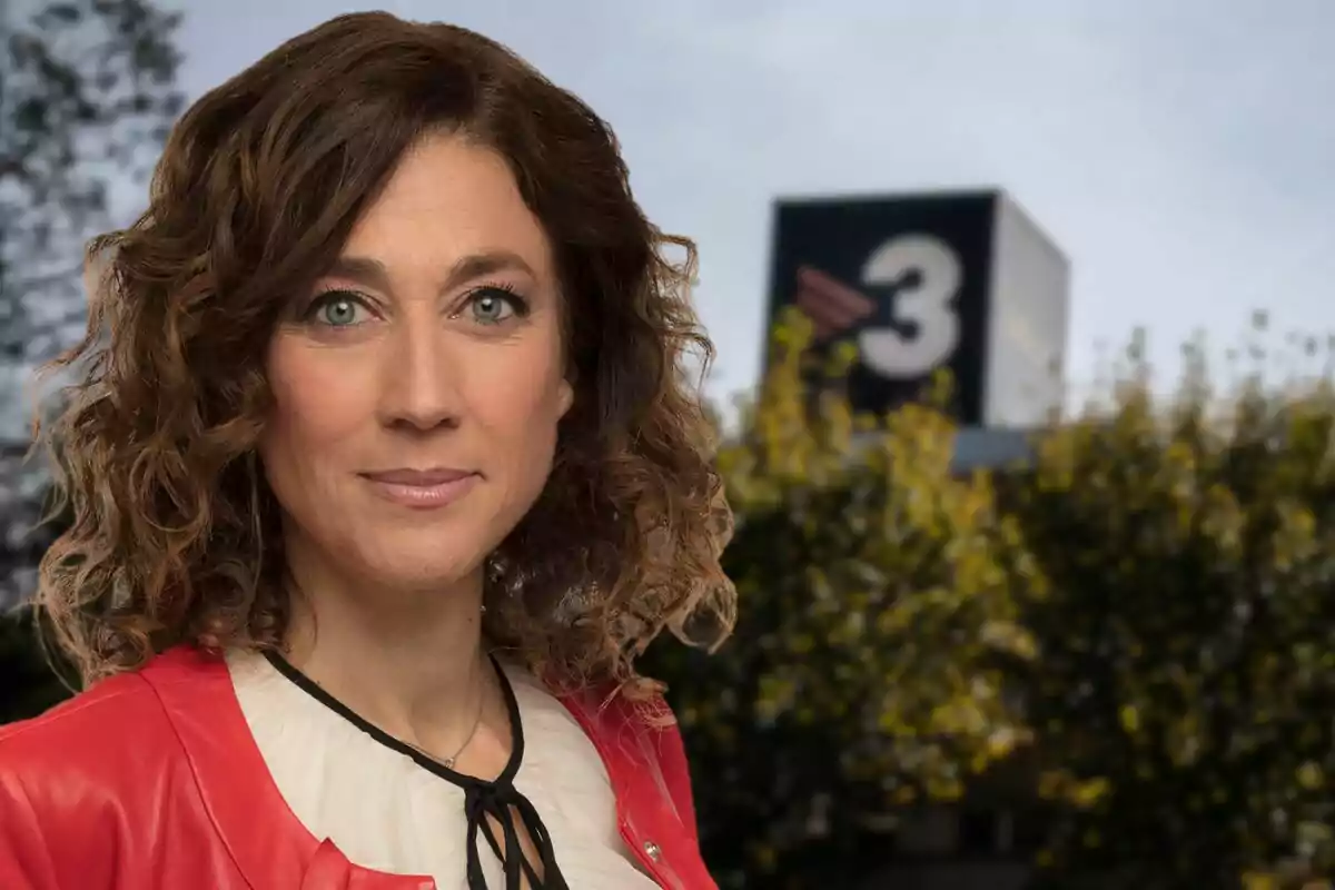 A woman with curly hair and a red jacket stands in front of a blurred background with a building that has a number 3 on it.