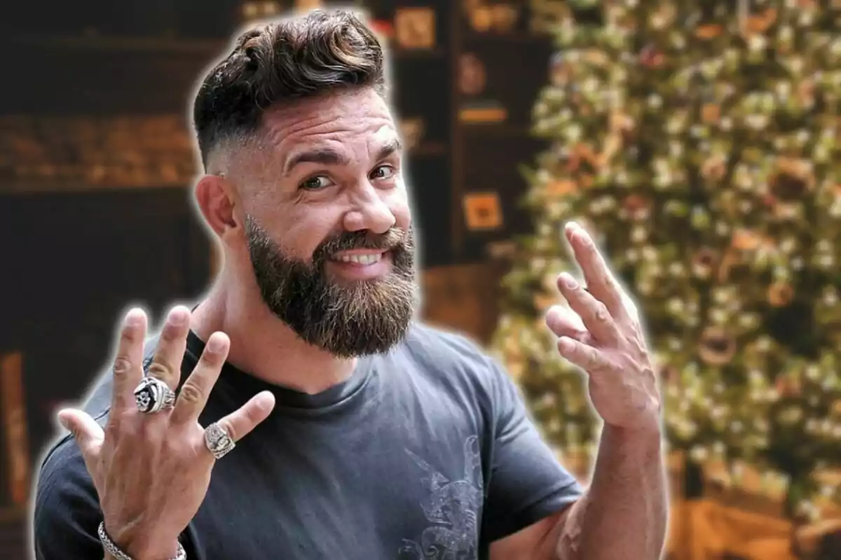 Hombre con barba y anillos sonríe mientras muestra sus manos frente a un árbol de Navidad desenfocado.