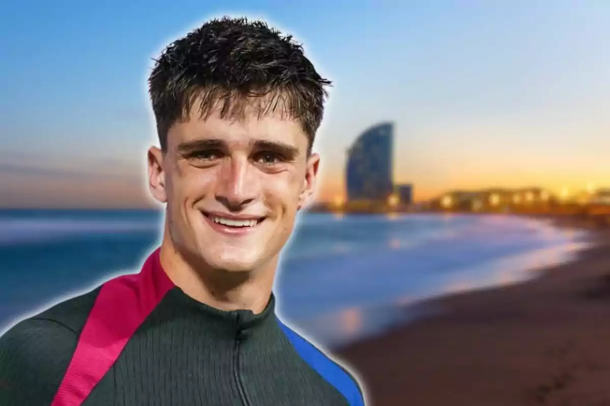 Un hombre sonriente con el cabello corto y oscuro lleva una chaqueta deportiva con detalles en rosa y azul con una playa y un edificio moderno al fondo al atardecer.