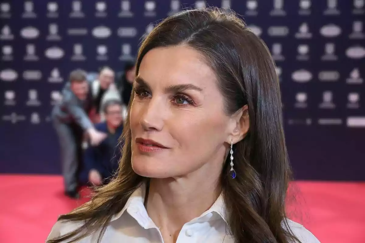 Una mujer con cabello castaño y aretes elegantes en un evento con alfombra roja y fondo de logotipos.