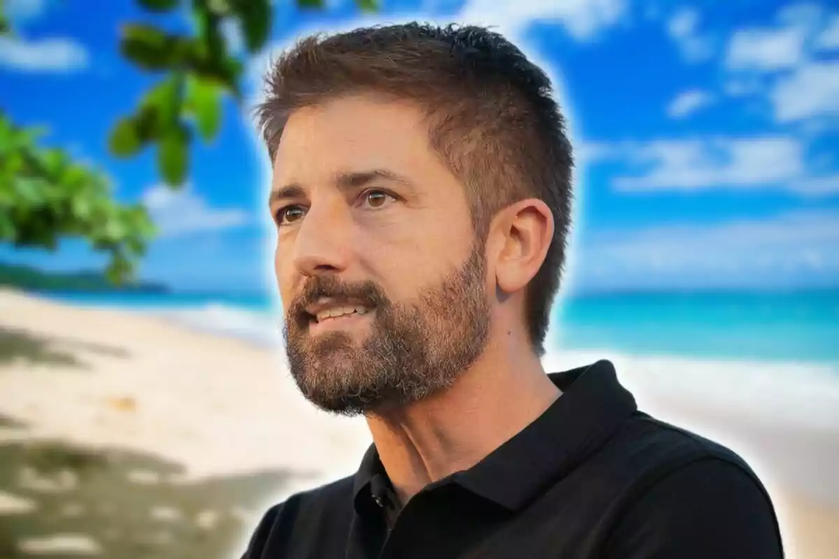 Un home amb barba i cabells curts mira cap a la dreta mentre està en una platja amb el mar i el cel blau de fons.
