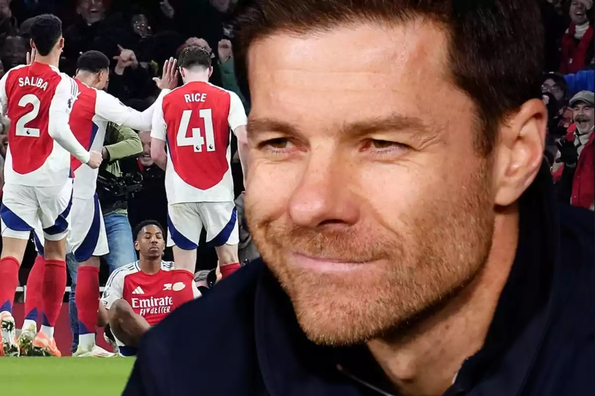 Close-up of a smiling man with Arsenal football players celebrating on the field in the background