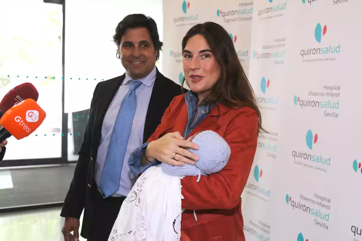 Lourdes Montes holds a baby wrapped in a blanket while Fran Rivera smiles beside her in front of a background with the Quirónsalud logo and several press microphones. Lourdes Montes holds a baby wrapped in a blanket while Fran Rivera smiles beside her in front of a background with the Quirónsalud logo and several press microphones.