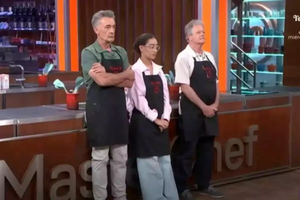 Francis Lorenzo, Marina Rivers i Juan Luis Cano amb davantals negres estan drets en una cuina de MasterChef Celebrity.