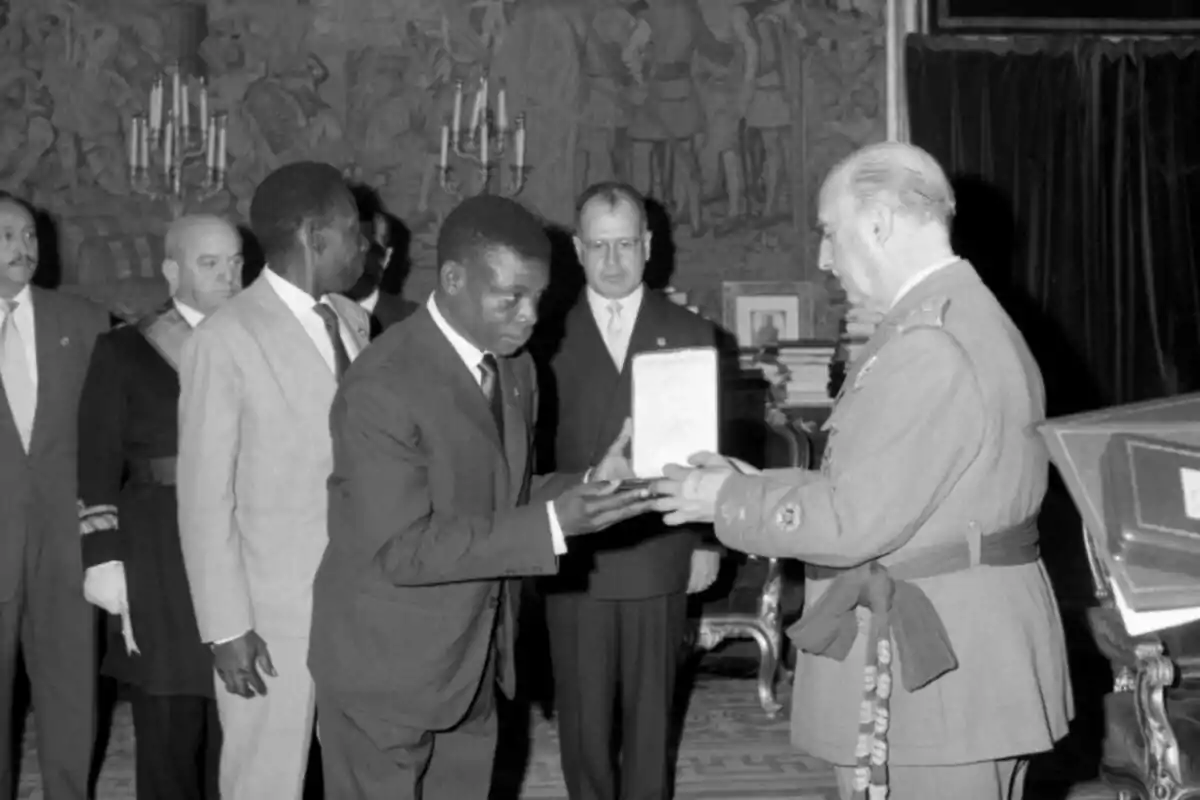 Un home amb vestit rep un document de mans d’un militar en una cerimònia formal amb diverses persones observant en un saló elegant. Un home amb vestit rep un document de mans d’un militar en una cerimònia formal amb diverses persones observant en un saló elegant.