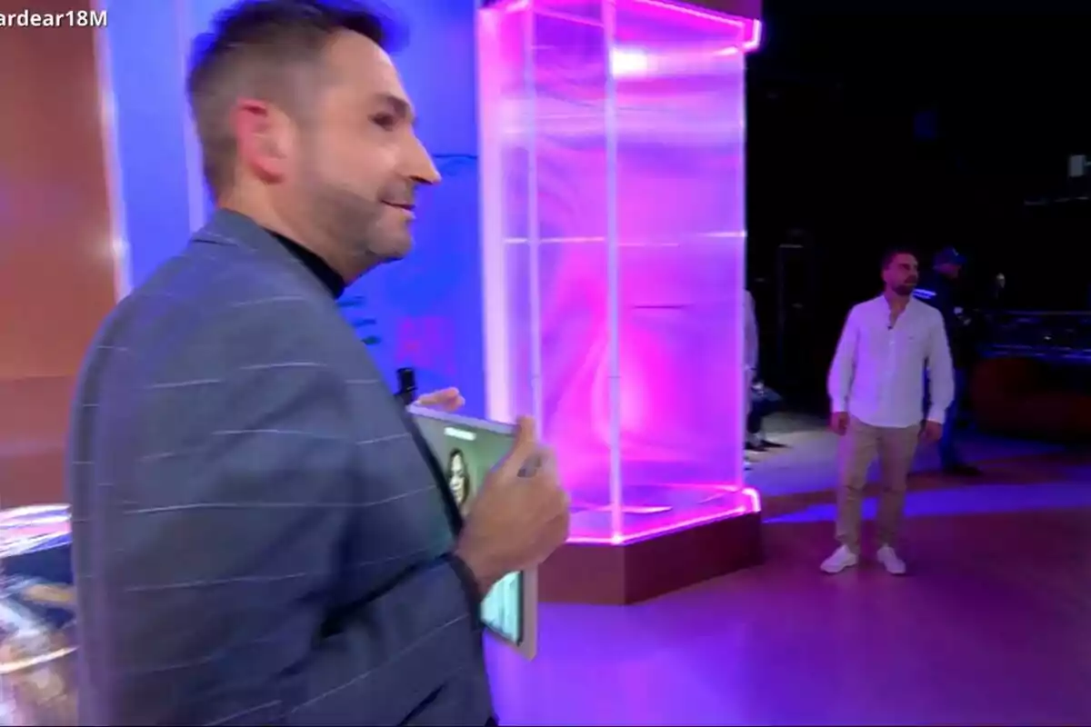 Frank Blanco amb jaqueta sosté una tauleta mentre camina en un estudi de Tardear il·luminat amb llums de colors, al fons es veu a José María Almoguera dret. Frank Blanco amb jaqueta sosté una tauleta mentre camina en un estudi de Tardear il·luminat amb llums de colors, al fons es veu a José María Almoguera dret.