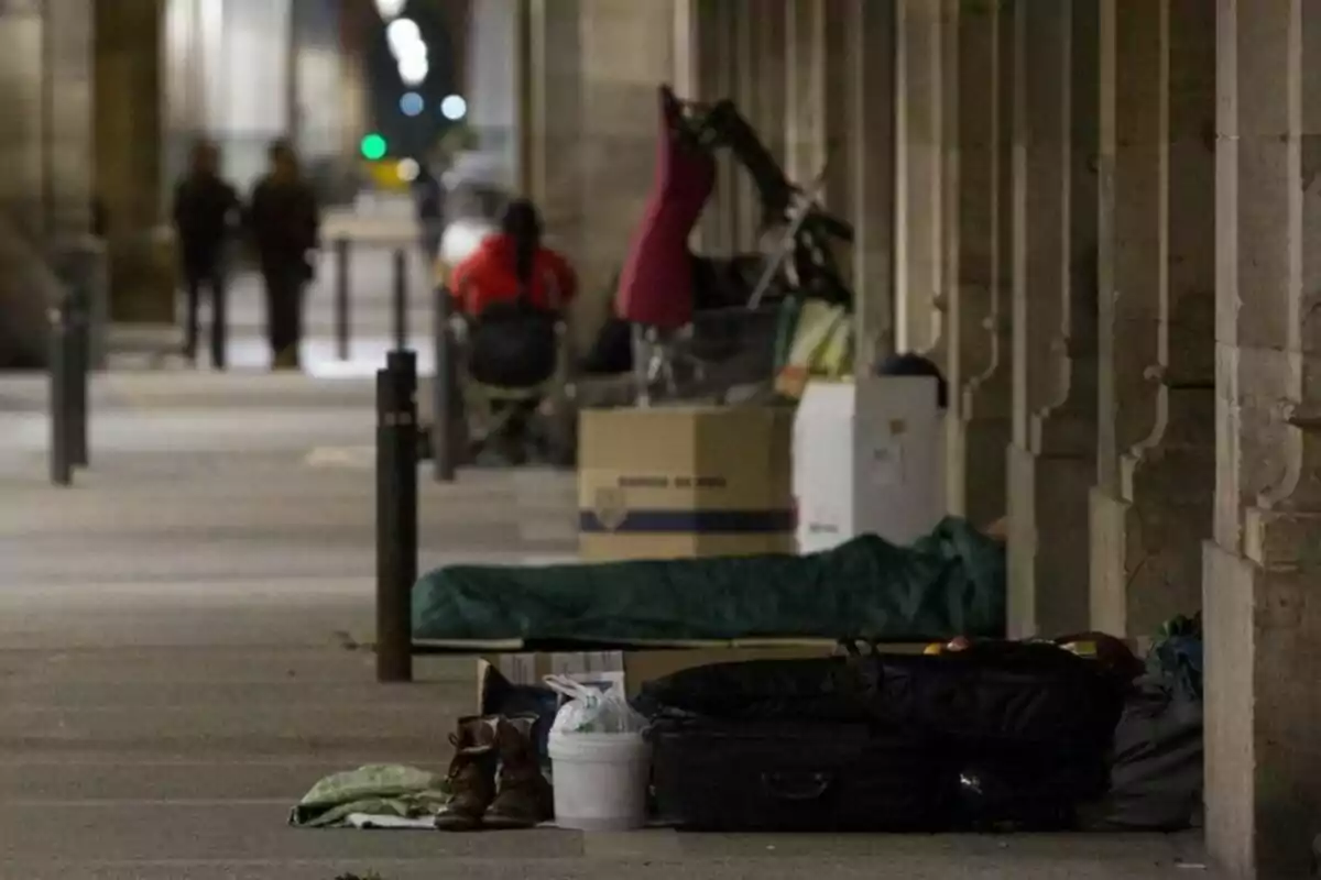 A hallway covered with homeless people and their belongings, including boxes, bags, and a sleeping bag, while two people walk in the background.