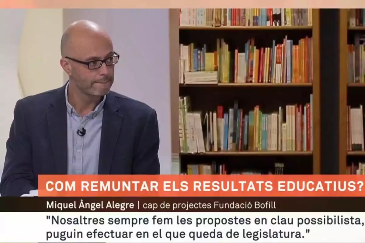 Un hombre con gafas y chaqueta oscura participa en un programa de televisión sobre educación, con una estantería llena de libros al fondo.