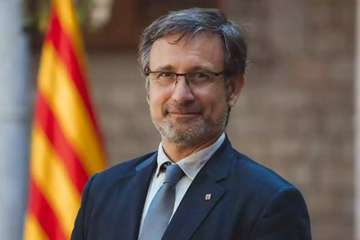 Un hombre con gafas y barba, vestido con traje y corbata, posa frente a una bandera con franjas rojas y amarillas.