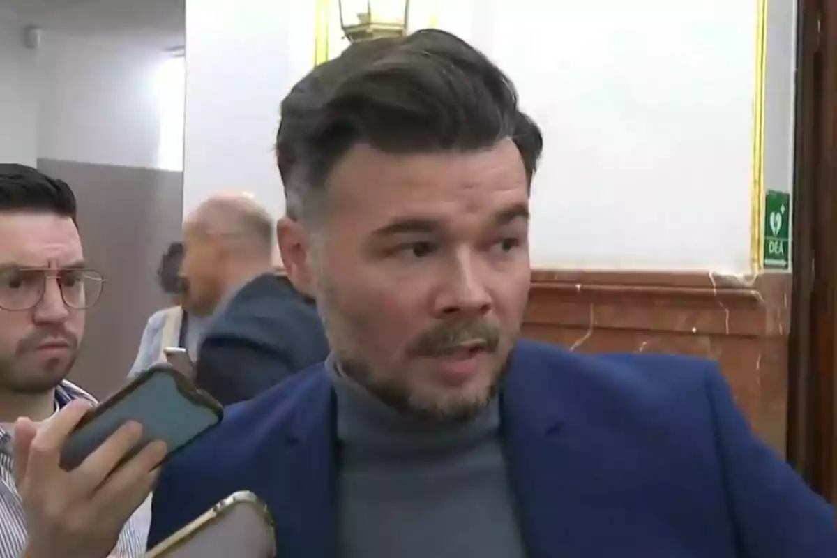 Un hombre con barba y cabello oscuro está hablando mientras otra persona sostiene un teléfono cerca de él en un entorno interior.