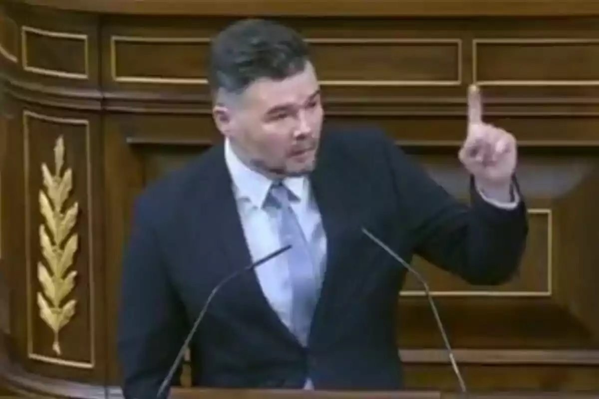Un home amb vestit i corbata està parlant en un faristol de fusta, aixecant un dit mentre s'adreça al públic. Un home amb vestit i corbata està parlant en un faristol de fusta, aixecant un dit mentre s'adreça al públic.