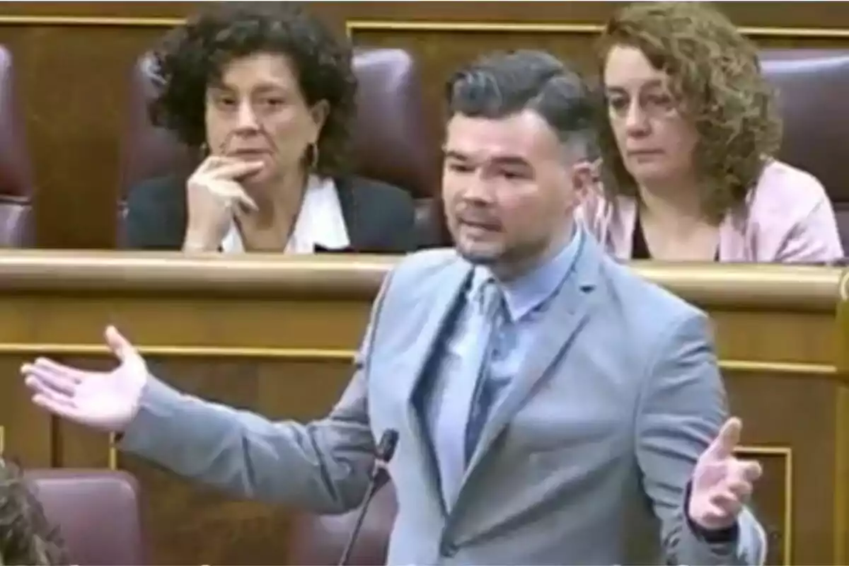Un home amb vestit gris gesticula mentre parla en un entorn parlamentari, amb dues dones assegudes darrere d'ell observant atentament. Un home amb vestit gris gesticula mentre parla en un entorn parlamentari, amb dues dones assegudes darrere d'ell observant atentament.