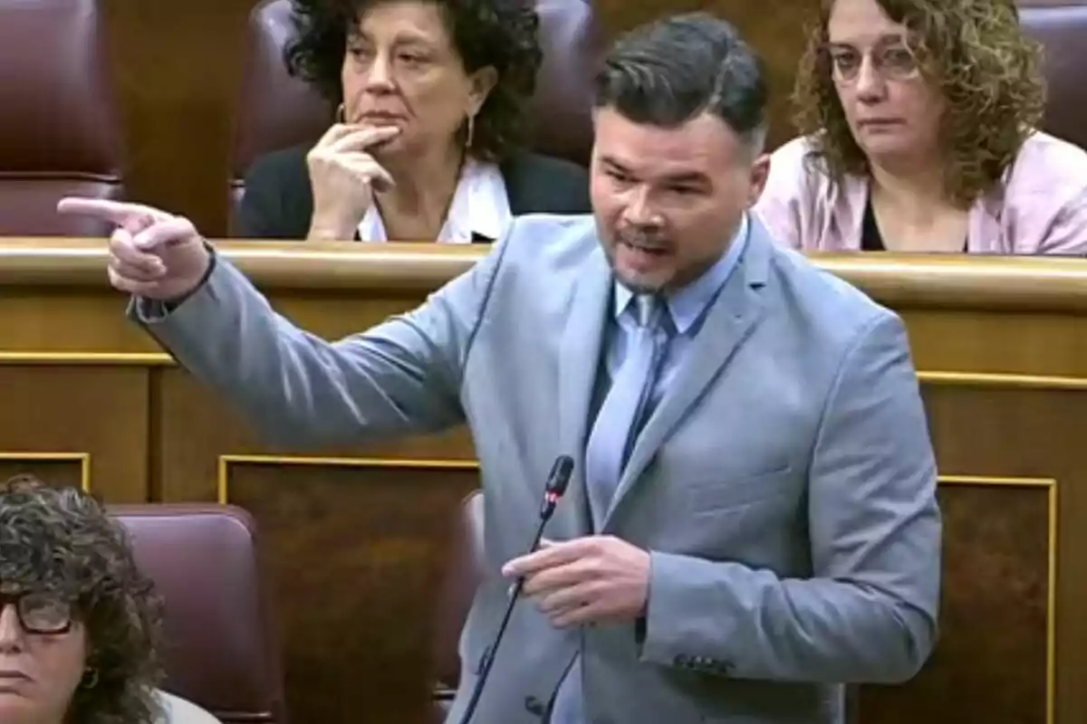 Un home amb vestit gris parla i gesticula en un parlament mentre altres persones l'observen des dels seus seients.