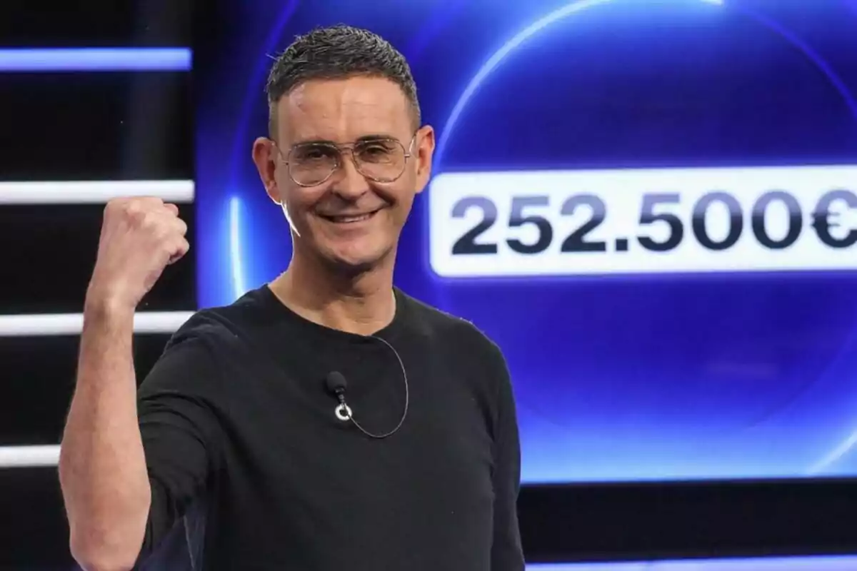 Home somrient amb ulleres i samarreta negra aixecant el puny en senyal de victòria davant d’un fons blau amb la xifra de 252.500 euros de Cifres i Lletres Home somrient amb ulleres i samarreta negra aixecant el puny en senyal de victòria davant d’un fons blau amb la xifra de 252.500 euros de Cifres i Lletres