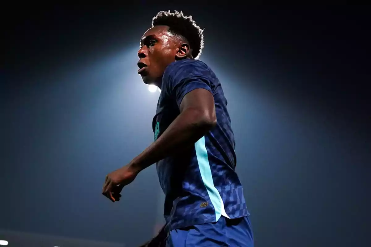 Un jugador de futbol amb uniforme blau fosc il·luminat per una llum de fons en un estadi.