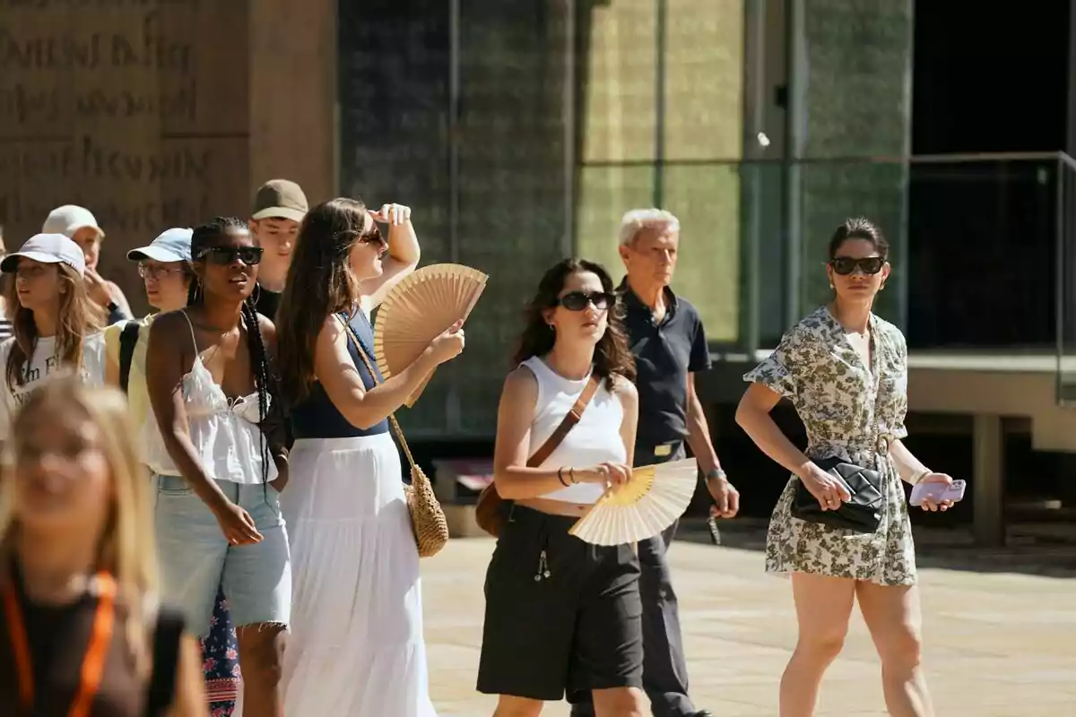 Grup de persones caminant sota el sol, algunes fent servir ventalls i ulleres de sol per protegir-se de la calor. Grup de persones caminant sota el sol, algunes fent servir ventalls i ulleres de sol per protegir-se de la calor.