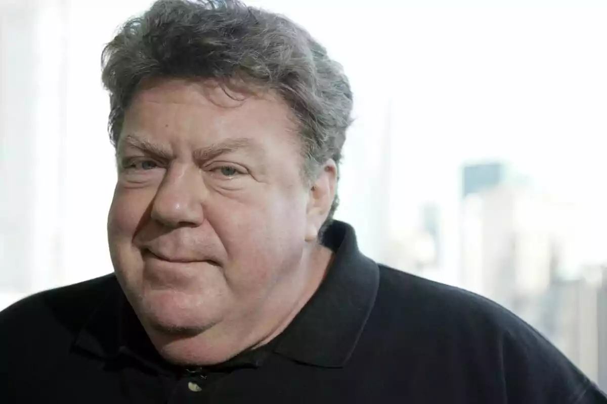 George Wendt amb cabell arrissat i una expressió neutral porta una camisa fosca amb un fons desenfocat. George Wendt amb cabell arrissat i una expressió neutral porta una camisa fosca amb un fons desenfocat.