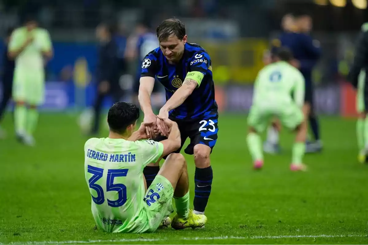 Un jugador de futbol amb uniforme blau ajuda a aixecar-se a un altre jugador amb uniforme verd que està assegut al camp. Un jugador de futbol amb uniforme blau ajuda a aixecar-se a un altre jugador amb uniforme verd que està assegut al camp.