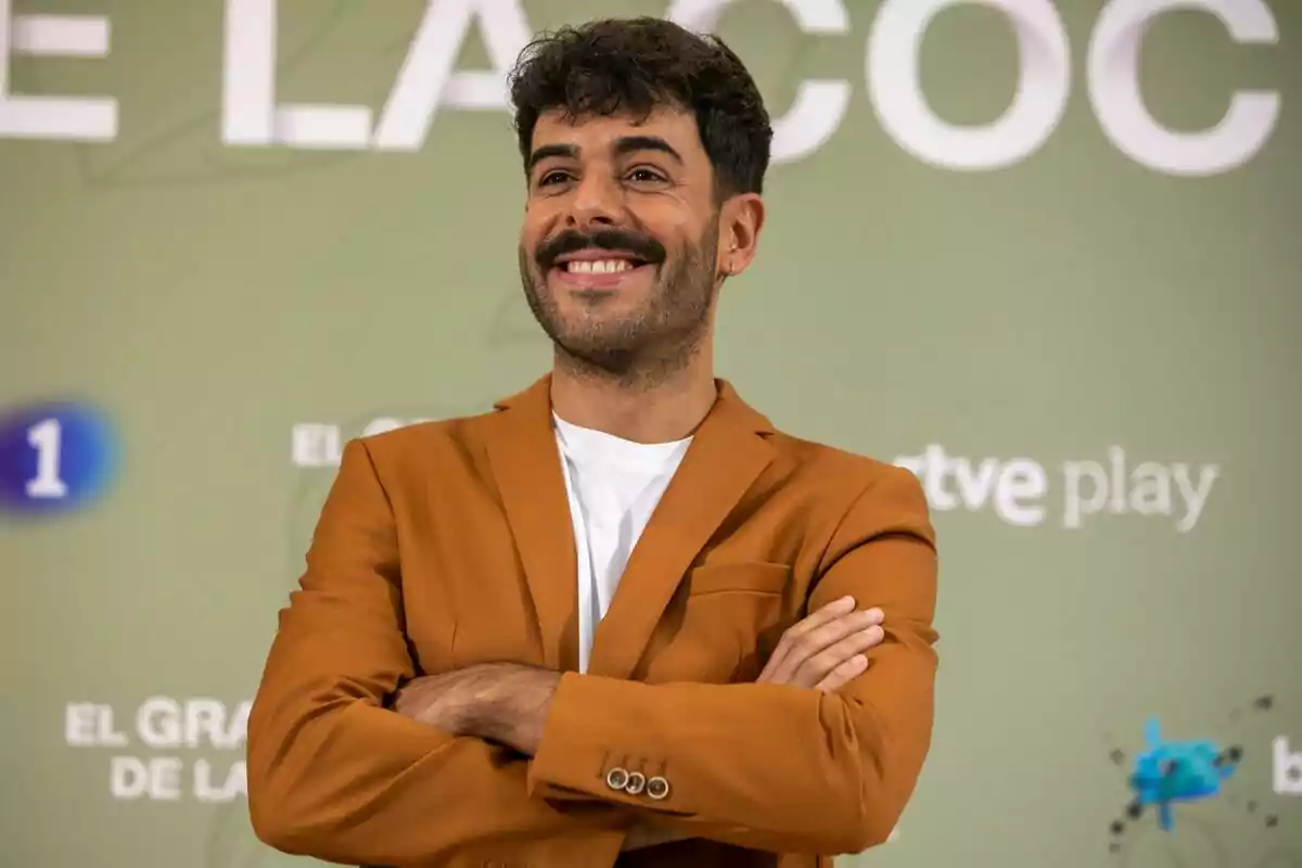 Germán González amb jaqueta marró i samarreta blanca somrient amb els braços plegats davant d'un fons verd amb text a la presentació del Gran Premi de la Cuina.