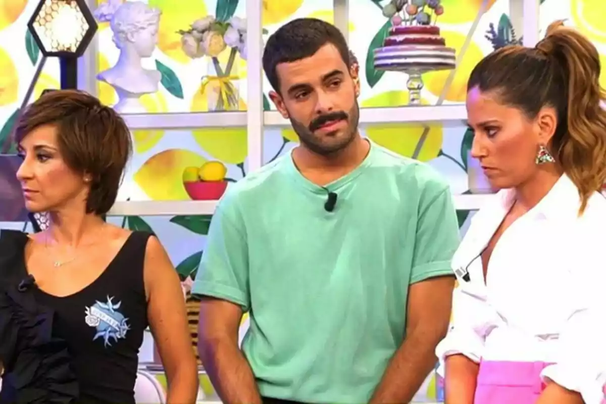 Adela González, Germán González i Nuria Marín estan drets en un set de televisió decorat amb motius de fruites i pastissos, una dona amb cabell curt i fosc porta una samarreta negra, un home amb barba i bigoti vesteix una samarreta verda, i una altra dona amb els cabells recollits porta una brusa blanca i una faldilla rosa al plató de Sálvame.