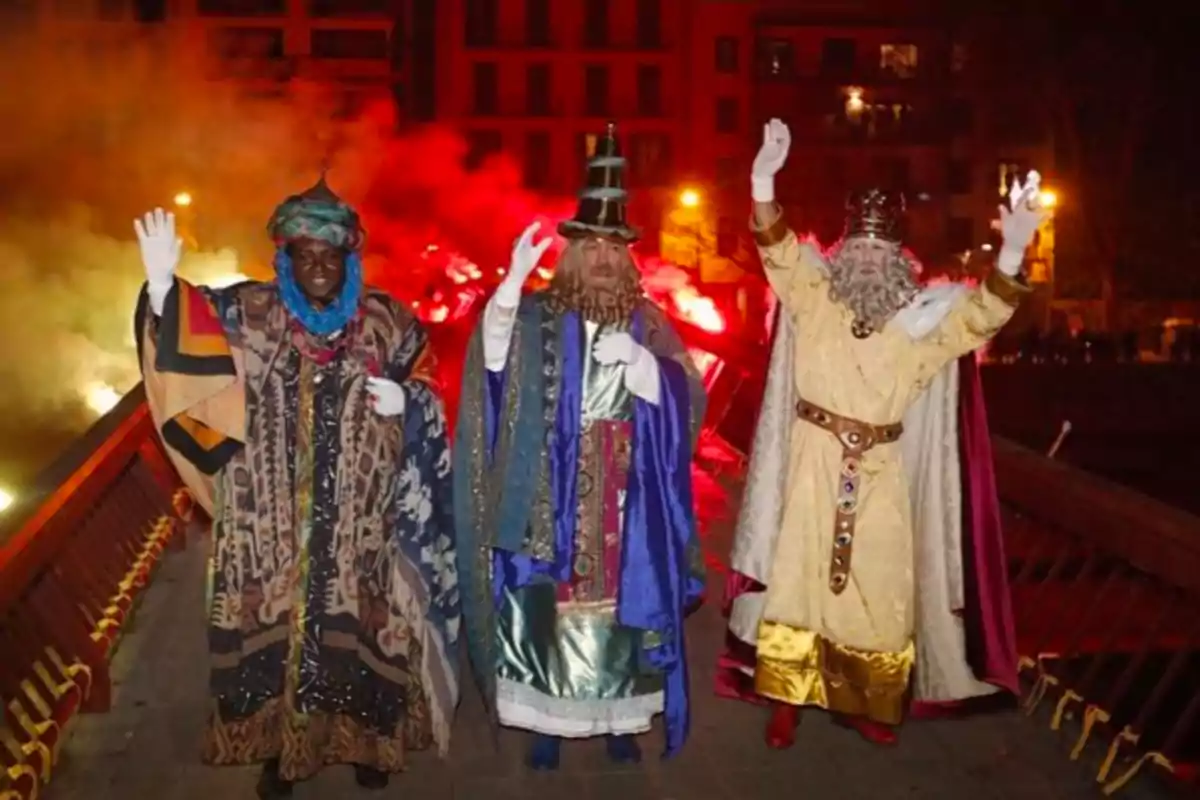 Tres persones disfressades de Reis Mags saluden mentre caminen per un pont il·luminat per llums vermelles al fons. Tres persones disfressades de Reis Mags saluden mentre caminen per un pont il·luminat per llums vermelles al fons.