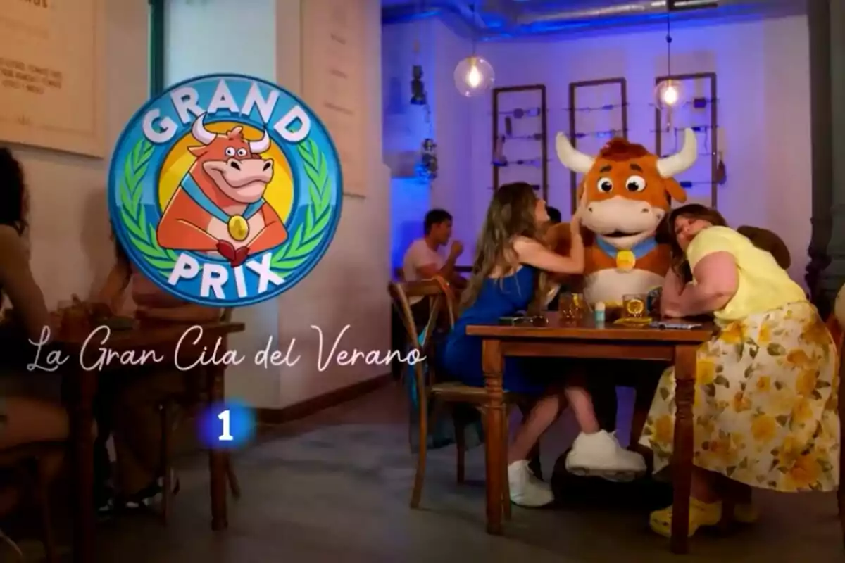 Lalachus i Ángela Fernández assegudes en una taula al costat d’una botarga de toro en un restaurant amb el logotip de Grand Prix i el text La Gran Cita de l’Estiu