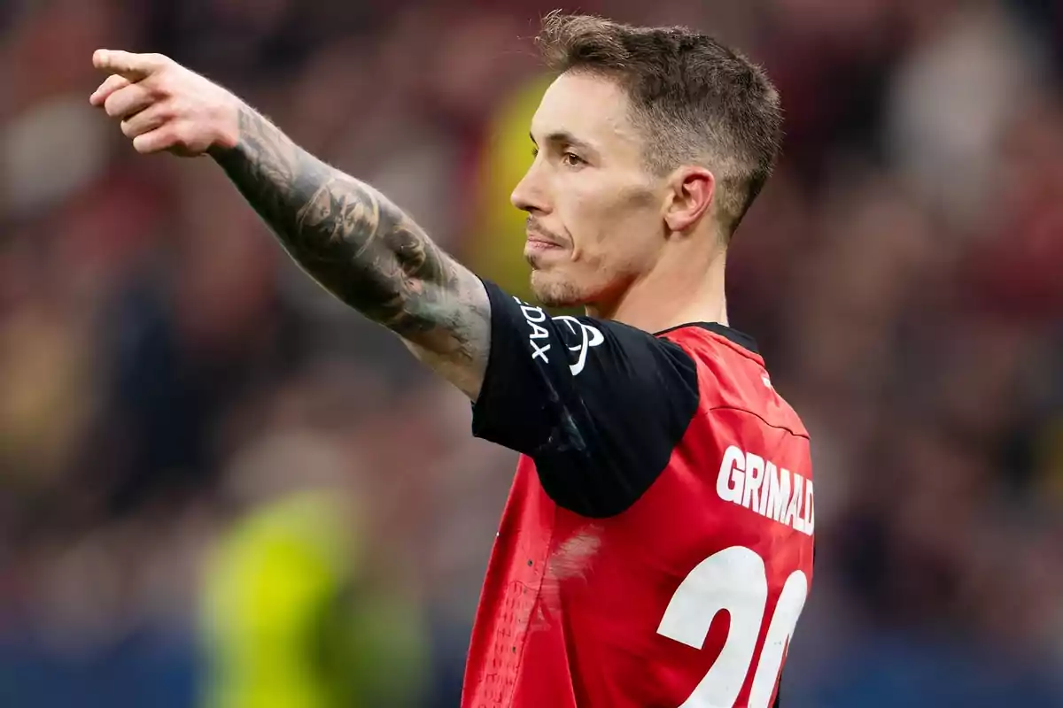 Un jugador de futbol amb tatuatges al braç assenyala cap endavant durant un partit, vestint una samarreta vermella amb el número 20. Un jugador de futbol amb tatuatges al braç assenyala cap endavant durant un partit, vestint una samarreta vermella amb el número 20.