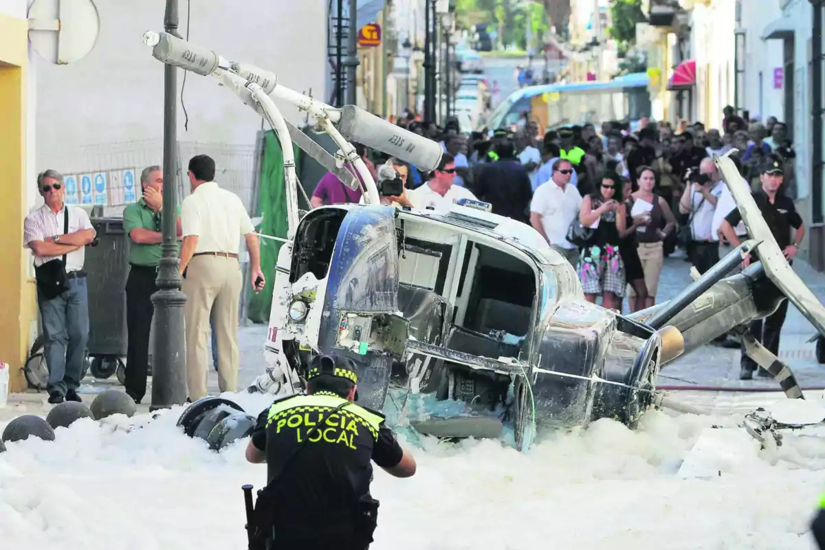 Un helicòpter accidentat jeu en un carrer envoltat d'escuma mentre un policia local observa l'escena i diverses persones s'agrupen al voltant. Un helicòpter accidentat jeu en un carrer envoltat d'escuma mentre un policia local observa l'escena i diverses persones s'agrupen al voltant.