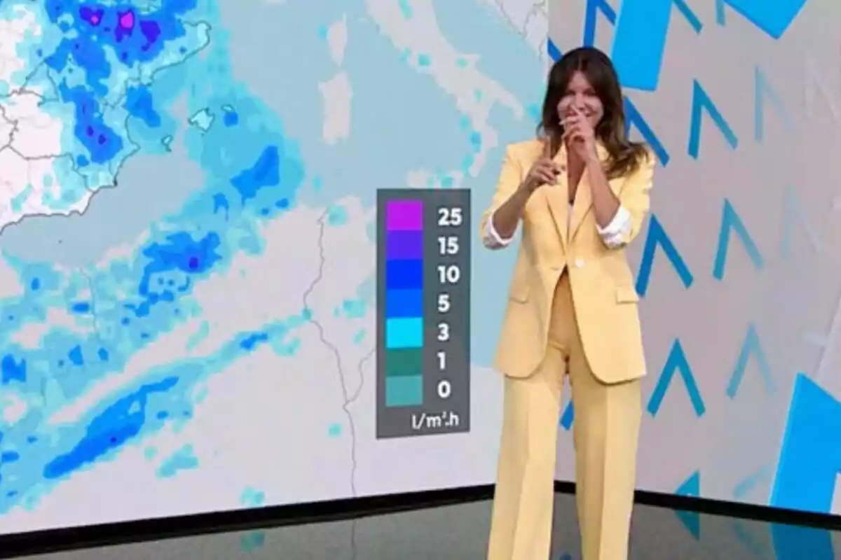 Himar González a Antena 3 Notícies, vestida amb un vestit groc, assenyala i somriu mentre mostra un mapa meteorològic amb diferents nivells de precipitació.