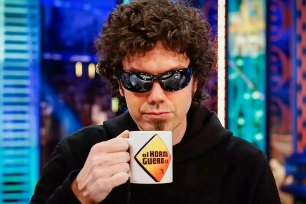 Pablo Ibáñez, El Hombre de Negro, con gafas de sol sostiene una taza con el logo de "El Hormiguero" en un ambiente colorido.