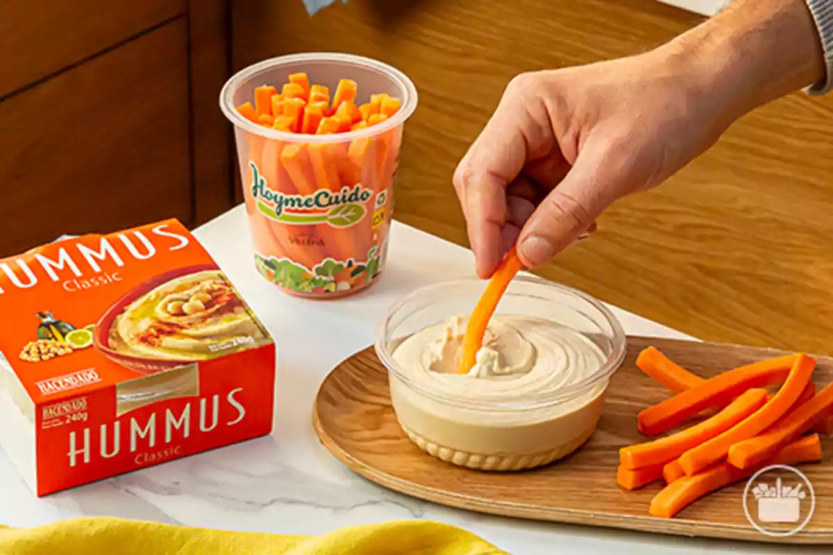 Una mà submergeix un bastonet de pastanaga en un recipient d’hummus al costat d’una safata amb més pastanagues i envasos d’hummus i pastanagues a la taula. Una mà submergeix un bastonet de pastanaga en un recipient d’hummus al costat d’una safata amb més pastanagues i envasos d’hummus i pastanagues a la taula.