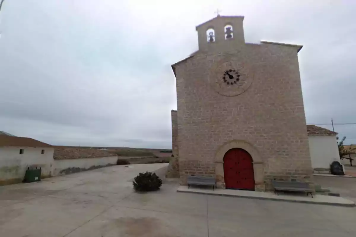 Una iglesia de piedra con un reloj en la fachada y una puerta roja, situada en un entorno rural con cielo nublado. Una iglesia de piedra con un reloj en la fachada y una puerta roja, situada en un entorno rural con cielo nublado.