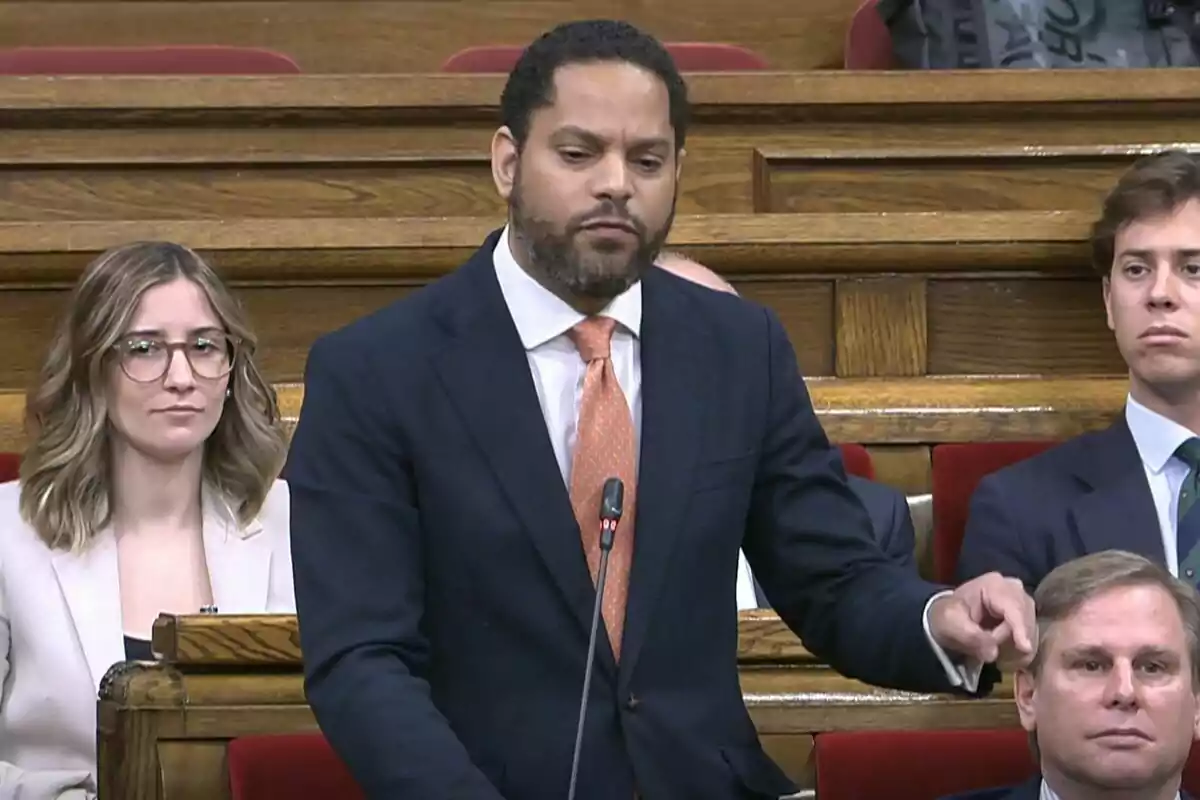 Un home dret en un entorn formal, possiblement un parlament, parlant mentre altres persones l'observen.