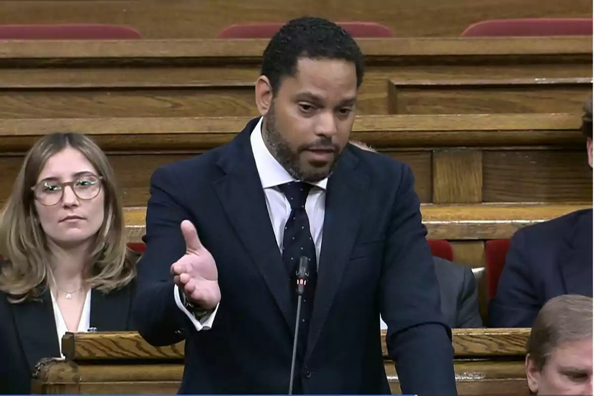Un home amb vestit fosc parla gesticulant amb la mà mentre està dret en un recinte parlamentari amb altres persones assegudes al seu voltant. Un home amb vestit fosc parla gesticulant amb la mà mentre està dret en un recinte parlamentari amb altres persones assegudes al seu voltant.
