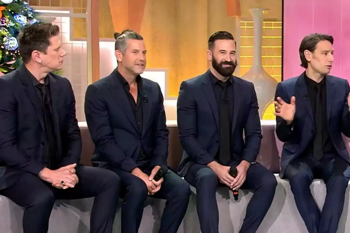 Quatre homes del grup Il Divo vestits amb vestits foscos estan asseguts en un sofà durant una entrevista en un set de televisió decorat amb un arbre de Nadal i gerros a Fiesta.