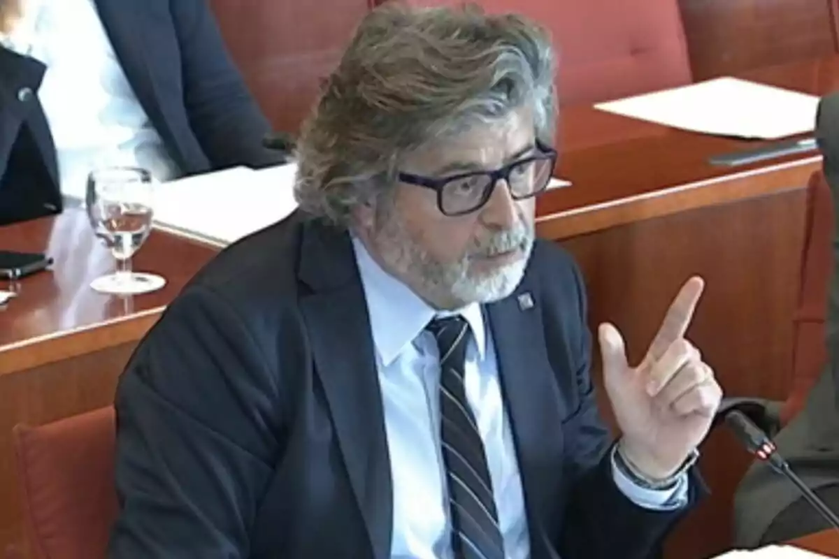 A man with a beard and glasses is speaking at a formal meeting while gesturing with his hand. A man with a beard and glasses is speaking at a formal meeting while gesturing with his hand.